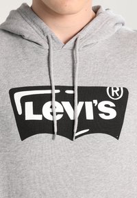 Persona que lleva una sudadera ligera gris con un gran logo en blanco y negro de Levi's en el pecho, mostrando los cordones de la sudadera colgando.