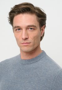 Blauer gestrickter Pullover mit Rundhalsausschnitt, der eine weiche Textur und ein leichtes Melange-Muster aufweist. Das Modell zeigt einen neutralen Gesichtsausdruck vor einem schlichten Hintergrund.