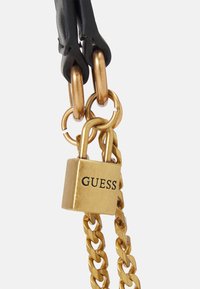 Candado dorado con el logo "GUESS", conectado a correas de cuero negro y eslabones de cadena dorada, exhibiendo un diseño elegante y acabado metálico.