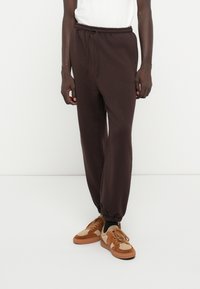 Braune Baumwoll-Jogginghosen mit einem elastischen Bund, seitlichen Taschen und schmal zulaufenden Bündchen, kombiniert mit braunen und beigen strukturierten Sneakers.