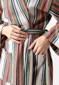 Accappatoio a righe in tonalità multicolori di rosso, verde, blu e toni neutri. Presenta una cintura, due tasche frontali e un tessuto strutturato.