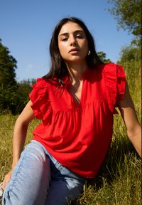 Rød kortærmet bluse med rynkede detaljer, v-udskæring og en afslappet pasform, samt lyseblå jeans i en naturlig indstilling.