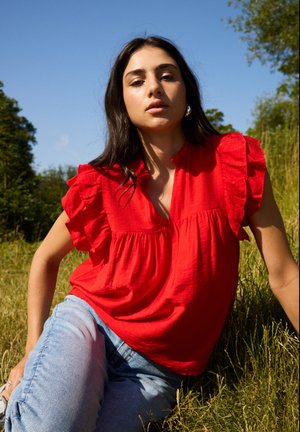 Rode blouse met korte mouwen, versierd met volants, een V-hals en een losse pasvorm, gecombineerd met lichtblauwe jeans in een natuurlijke omgeving.