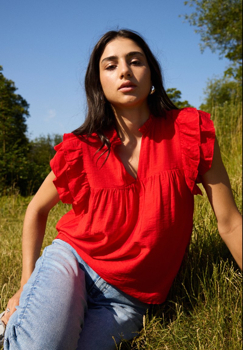 Rød kortærmet bluse med rynkede detaljer, v-udskæring og en afslappet pasform, samt lyseblå jeans i en naturlig indstilling.