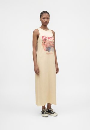 Frau trägt ein ärmelloses beiges Maxikleid mit dem Aufdruck „Le Riviera“ und schwarzen Sneakers, steht vor einem schlichten weißen Hintergrund.