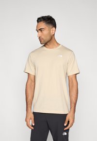 Μπεζ κοντομάνικο μπλουζάκι με στρογγυλή λαιμόκοψη και λευκό λογότυπο στο αριστερό στήθος. Το ύφασμα φαίνεται μαλακό με λεία υφή.