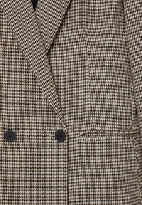 Blazer à motif vichy marron et beige avec un design à double boutonnage, comportant deux boutons noirs et une poche poitrine.
