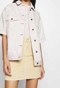 Giacca di jeans beige chiaro con maniche corte, tasche sul petto e bottoni marroni, abbinata a una gonna in velluto a coste giallo chiaro.