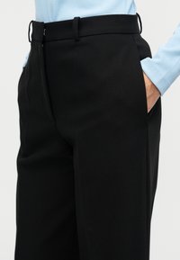 Μαύρο, ραμμένο παντελόνι με λεία υφή, που διαθέτει ψηλή μέση, πλαϊνές τσέπες και θηλιές για ζώνη. Συνδυασμένο με ανοιχτό μπλε τοπ από πάνω.