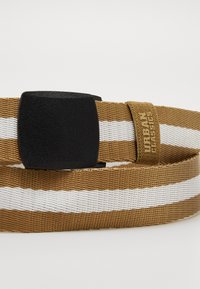Cinturón trenzado en marrón con franjas blancas, con una hebilla rectangular negra y una etiqueta en color marrón claro que dice "Urban Classics."