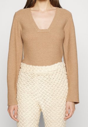 Femme portant un pull col V à manches longues couleur beige et un pantalon crème texturé taille haute avec un motif tricoté.