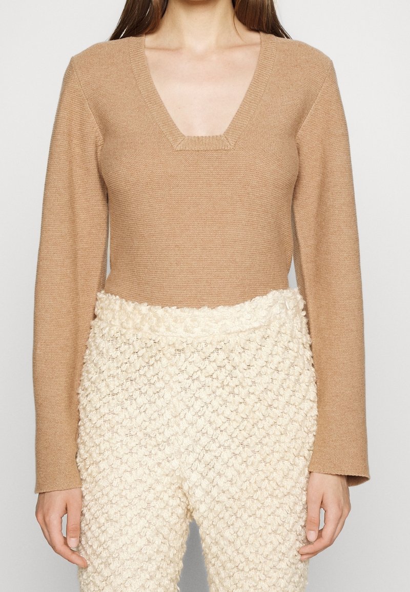 Femme portant un pull col V à manches longues couleur beige et un pantalon crème texturé taille haute avec un motif tricoté.