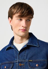 Džínová bunda v tmavě modré barvě s špičatým límcem, dvojitými náprsními kapsami a žlutými švy. Obsahuje značkové detaily s kovovými knoflíky.