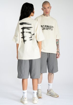Oversized cream t-shirt with black abstract print on back, paired with gray shorts. Model's shoes are beige with accents of white.