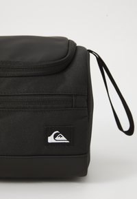 Bolsa aislante negra con una superficie texturizada, cierre de cremallera y un asa lateral. Cuenta con un parche de logo blanco en el frente.