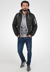 Sort zip-up jakke med hætte, med hvide snore og et logo på brystet. Bæret over en grå sweatshirt og parret med blå jeans og sneakers.