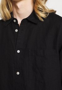 Černá košile na knoflíky vyrobená z texturované látky, s kapsou, špičatým límcem a bílými knoflíky, zdůrazňující jednoduchý design.