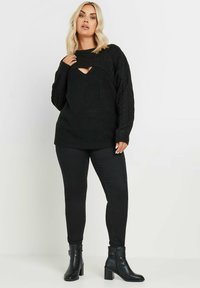 Mustat neuleet, joissa on leikkauksia rinnassa, yhdistettynä mustiin tiukkoihin housuihin ja nilkkureihin, joissa on paksut korot.