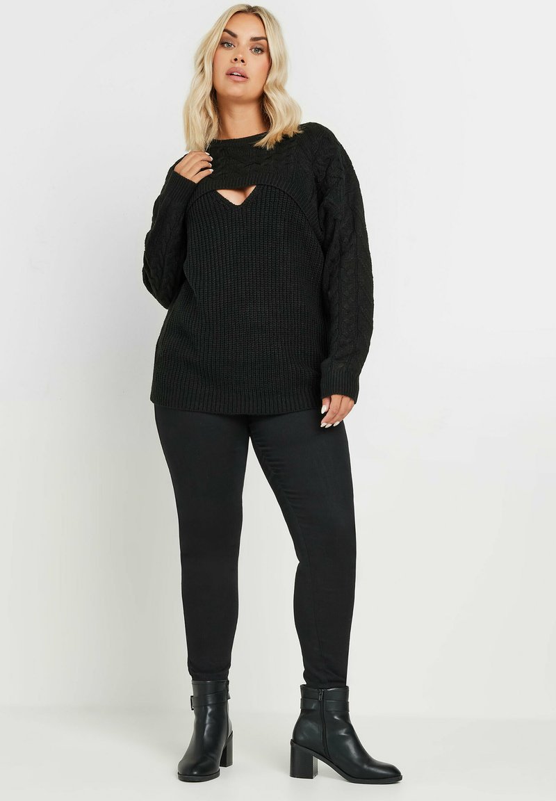 Mustat neuleet, joissa on leikkauksia rinnassa, yhdistettynä mustiin tiukkoihin housuihin ja nilkkureihin, joissa on paksut korot.