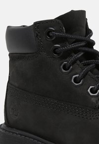 Bottine en cuir noir avec un bout rond, des lacets noirs contrastants, une texture lisse et des œillets en métal. Présente un logo sur le côté.