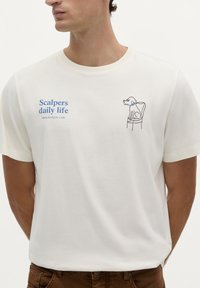 Weißes Baumwoll-T-Shirt mit blauer Aufschrift "Scalpers daily life" und einer Illustration eines Hundes, der auf einem Stuhl sitzt. Kurze Ärmel, runder Halsausschnitt.