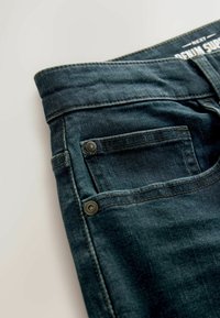 Jeans de denim albastru închis, cu o textură moale, dotate cu un buzunar mic, peițe de cupru și cusături vizibile pe talie și pe cusături.