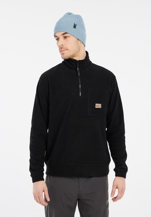 Čierny fleece pulóver s polovičným zipsom, s predným vreckom a logom na nášivke. Nosí sa s svetlomodrou pletenou čiapkou.