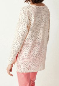 Haut en dentelle blanche avec motifs ajourés décoratifs, manches longues et encolure arrondie, associé à un pantalon rose clair.