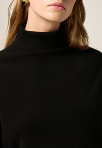 Maglione a collo alto di colore nero realizzato in tessuto morbido, caratterizzato da un design aderente con texture a costine vicino al collo e vista ravvicinata della parte superiore del corpo.