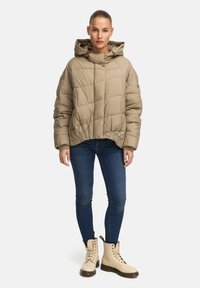 Beige pufferjack met horizontale stiksels, oversized capuchon en elastische zoom. Gecombineerd met aansluitende jeans en crèmekleurige veterschoenen.