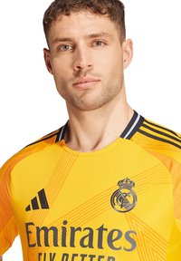 Žuta golmanska majica s crnim akcentima i prugama. Sadrži logo "Emirates" i grb Real Madrida na prsima. Glatka tekstura.