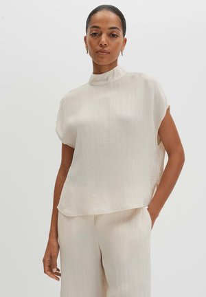 Vrouw met donker haar in een hoge knot, draagt een losse, gestructureerde witte top met korte mouwen en bijpassende wijde broek, hand in zak.