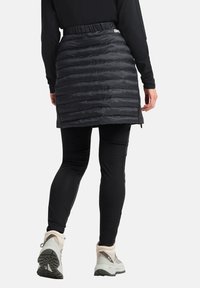 Zwarte pufferrok met een gewatteerd ontwerp, elastische taille en zijsplitten, gecombineerd met aansluitende zwarte leggings en lichtgekleurde wandelschoenen.