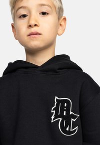 Sudadera negra de tejido suave con un logo blanco bordado texturizado en la parte izquierda del pecho. El diseño incluye una capucha amplia.