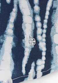 Marineblå stof med hvide abstrakte mønstre og en sølvcharms med et cirkulært design med udskæringer, der viser intrikate detaljer og tekstur.