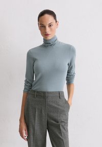 Frau mit dunklen Haaren in einem hellblauen Rollkragenpullover und grauen Nadelstreifen-Hosen, die vor einer einfachen weißen Wand steht.