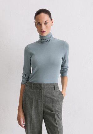 Donna con capelli scuri, in un maglione a collo alto blu chiaro e pantaloni grigio a righe, in piedi contro un muro bianco semplice.