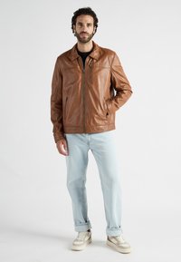 Man in een bruine leren jas, lichtblauwe jeans en witte sneakers, staande met één hand in de zak tegen een effen achtergrond.