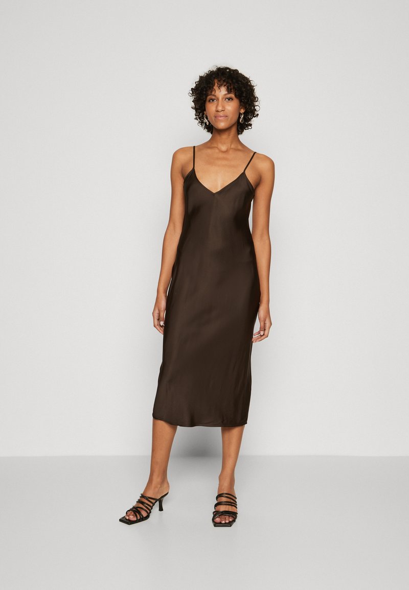 Robe nuisette en satin marron avec un décolleté en V, des bretelles fines et ajustables, et une longueur mi-mollet. Associée à des sandales à talons compensés noires.