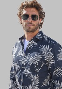 Marineblauw overhemd met witte palmenpatroon, voorzak en met kraag. Model draagt een zonnebril en een wit T-shirt eronder.