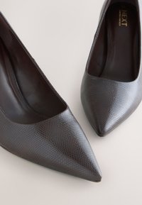 Escarpins en cuir marron foncé à bout pointu avec un motif texturé subtil, étiquetés "NEXT" à l'intérieur, posés sur une surface claire.
