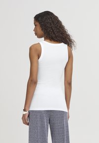 Vrouw met krullend haar, gekleed in een witte mouwloze top en wijde broek met patroon, staand met haar rug naar de camera tegen een effen achtergrond.