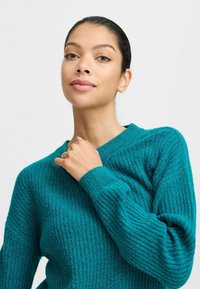 Pull en maille côtelée bleu sarcelle avec un col en V et des manches longues, présentant un tissu texturé et une coupe décontractée. Gros plan sur la main au niveau de l'encolure.