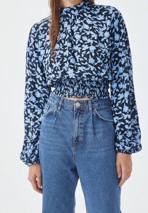 Woman wearing long-sleeve navy blouse with blue floral pattern and smocked waist paired with high-waisted blue denim jeans.