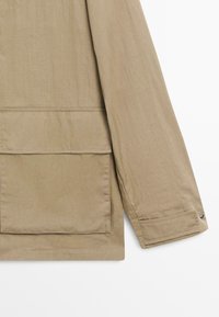 Beige jas mouw met grote voorzak en geribbelde manchet met knoopdetail op een witte achtergrond.