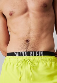 Heldergele zwembroek met een zwarte elastische tailleband met daarop "CALVIN KLEIN" in het wit, en een trekkoordsluiting. Soepele textuur.