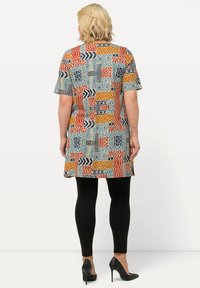 Monivärinen tunika, jossa on abstrakteja geometrisia kuvioita punaisena, sinisenä ja oranssina vaalealla taustalla, yhdistettynä mustiin legginsseihin ja korkokenkiin.