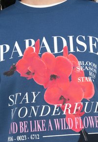 Μπλε βαμβακερό T-shirt με μεγάλα κόκκινα γραφικά ορχιδέας και λευκό και ροζ κείμενο. Κοντά μανίκια και στρογγυλή λαιμόκοψη.