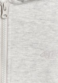 Gros plan d'un tissu gris clair avec une fermeture éclair blanche et un logo "4F" brodé discret sur le côté droit.