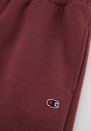 Pantalones de chándal burdeos hechos de un material suave, con una cintura elástica, bolsillos laterales y un pequeño logotipo bordado en la parte frontal.